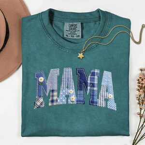 以下が含まれることがあります： 「MAMA」の文字がパッチワーク生地でアップリケされた、ティール色のComfort Colors Tシャツ。文字は様々な青と白の色合いで、デイジーのアクセントが付いています。金の星のネックレスがシャツの上にかけられています。