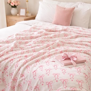 May include: A bed is covered with a white blanket patterned with pink bows and hearts. A pink gift box with a matching bow sits on top of the blanket. A pink pillow rests against white pillows.