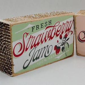 Puede incluir: Dos letreros de madera con un diseño rústico. Uno dice "FRESH Strawberry Jamo" en letras rojas y blancas sobre un fondo verde. El otro letrero dice "the Strawberry Patch" en letras rojas sobre un fondo rosa.