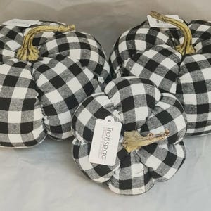 May include: Three fabric pumpkins featuring a black and white buffalo check pattern. Each pumpkin has a textured, light-colored stem at the top. One smaller pumpkin in the front displays a white rectangular tag with the text Transpac.