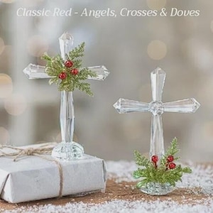 May include: Two clear glass crosses, one atop a wrapped gift, the other on a snowy surface. The crosses are decorated with green sprigs and red berries. The text "Classic Red - Angels, Crosses & Doves" is at the top.