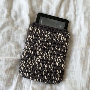 May include: A rectangular crochet sleeve made of thick dark charcoal and cream-colored yarn, featuring a textured, interlocking stitch pattern. A black electronic e-reader device is partially inserted into the top opening of the protective pouch.