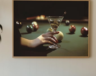 May include: A framed print shows a martini glass with olives on a pool table, with a hand holding the glass. Various coloured billiard balls are scattered on the green felt. The print is displayed above a brown tufted sofa with a yellow cushion.