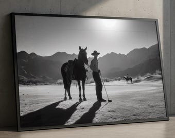 May include: Black and white framed print of a cowboy with a horse on a golf course. The cowboy is wearing a hat and holding a golf club. Mountains are in the background. The image is displayed against a concrete wall.