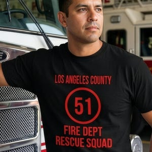 May include: Black t-shirt with red text that reads "LOS ANGELES COUNTY 51 FIRE DEPT RESCUE SQUAD." The shirt is worn by a person standing in front of a fire truck. The fire truck is red and white.