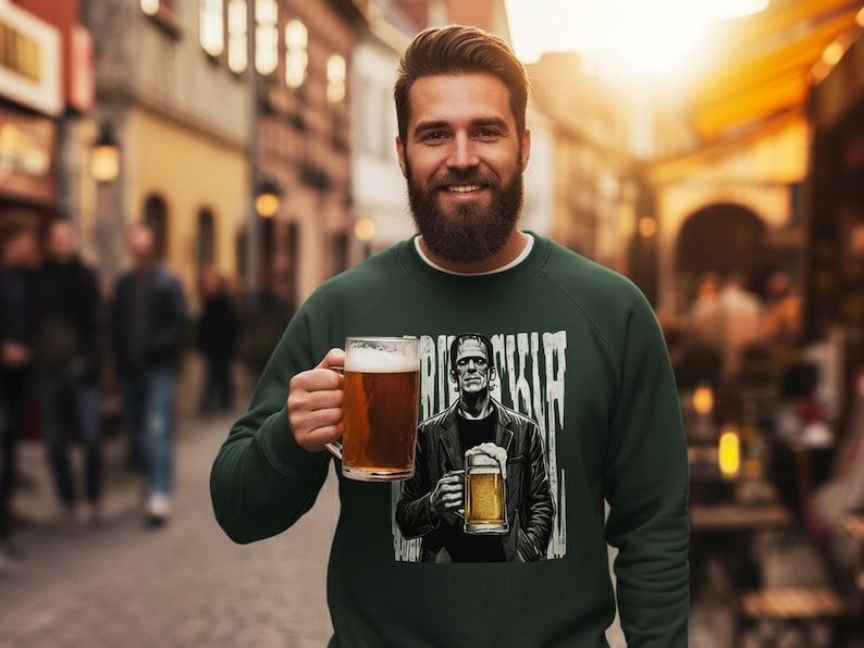 May include: A dark green long-sleeve sweatshirt featuring a graphic print of a character resembling Frankenstein's monster holding a beer mug, with the text "FRANKENSTEIN" partially visible in a distressed white font above the character.