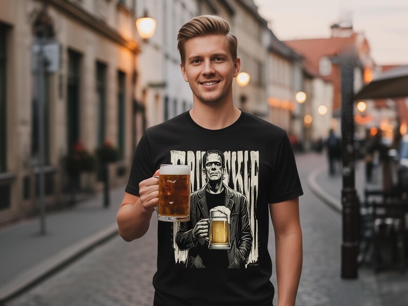 May include: A black t-shirt featuring a graphic print of a figure resembling Frankenstein's monster holding a beer mug, with the text "FRANKENSTEIN" displayed above the figure in a distressed, vertical font style, set against a light-coloured background.