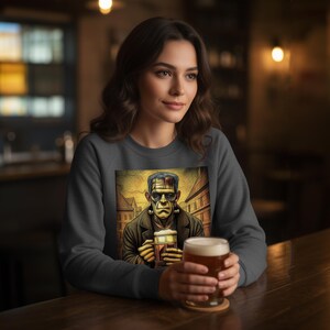 May include: A dark gray crewneck sweatshirt featuring a graphic print of a green-skinned monster with dark hair and neck bolts, holding a glass of beer. The illustration depicts the character in a city street setting with a warm color palette.