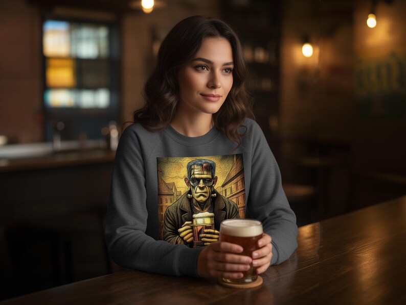 May include: A dark gray crewneck sweatshirt featuring a graphic print of a green-skinned monster with dark hair and neck bolts, holding a glass of beer. The illustration depicts the character in a city street setting with a warm color palette.