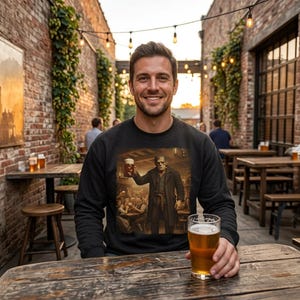 May include: A black long-sleeve sweatshirt featuring a graphic print of a figure resembling Frankenstein's monster holding a beer mug in a tavern setting. The sweatshirt is worn by a person sitting at a wooden table with a glass of beer.