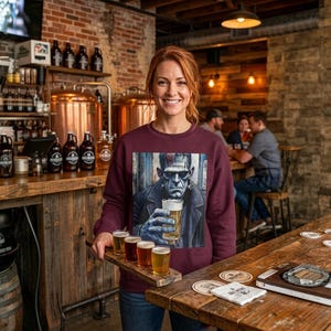 Pode incluir: Uma camisola de manga comprida cor de vinho com uma impress&atilde;o gr&aacute;fica do monstro de Frankenstein a segurar um copo de cerveja. A camisola &eacute; usada por uma pessoa que segura uma t&aacute;bua de madeira com quatro copos pequenos de cerveja numa cervejaria.
