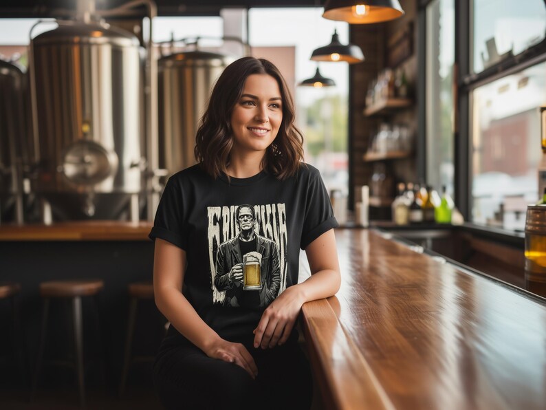 May include: A black short-sleeve t-shirt featuring a graphic print of a figure resembling Frankenstein's monster wearing a jacket and holding a glass of beer, with the text "FRANKENSTEIN" printed in a distressed, vertical font above the figure.