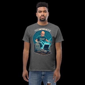 May include: Dark gray t-shirt with a graphic of a bald man with glasses, lightning bolts, a laptop, and a tombstone that says "RIP". The shirt has the word "TECHNOMANCER" in a circle above the graphic.