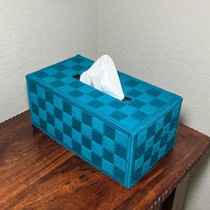 May include: A rectangular tissue box cover in teal and dark teal checkered pattern. White tissues are visible through the rectangular opening. The box is placed on a wooden surface.