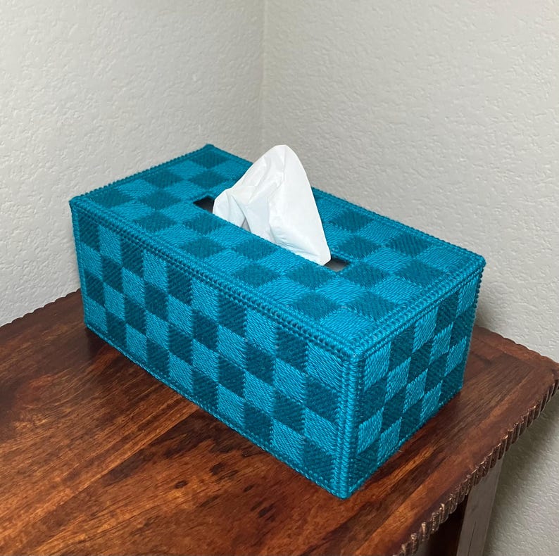 May include: A rectangular tissue box cover in teal and dark teal checkered pattern. White tissues are visible through the rectangular opening. The box is placed on a wooden surface.
