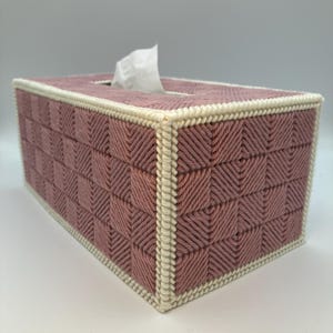 May include: A rectangular tissue box cover in a woven pattern. The box is primarily a dusty rose color with a cream-colored border. White tissues are visible extending from the top opening. The woven design creates a textured, geometric appearance.