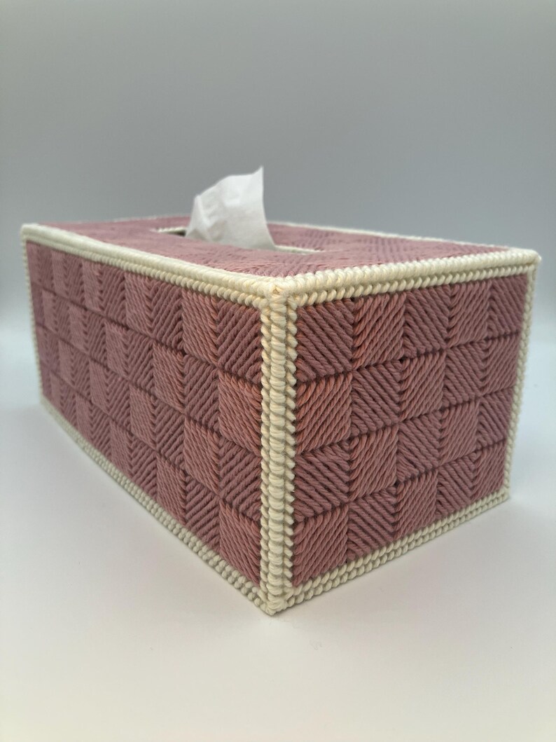 May include: A rectangular tissue box cover in a woven pattern. The box is primarily a dusty rose color with a cream-colored border. White tissues are visible extending from the top opening. The woven design creates a textured, geometric appearance.
