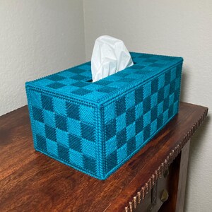 May include: A rectangular tissue box cover in a teal and dark teal checkerboard pattern. White tissues are visible from the top opening. The box is made of a woven material and sits on a wooden surface.