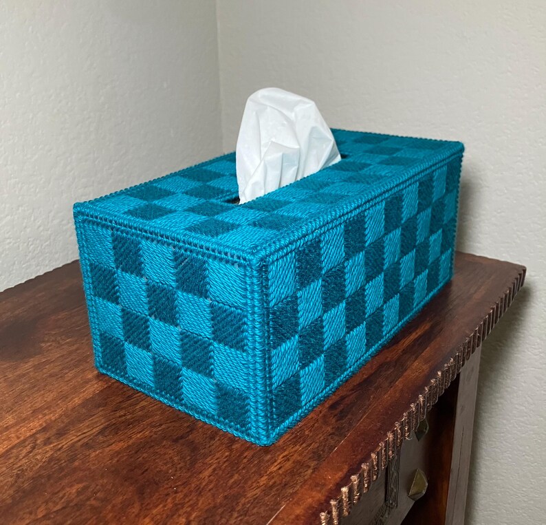May include: A rectangular tissue box cover in a teal and dark teal checkerboard pattern. White tissues are visible from the top opening. The box is made of a woven material and sits on a wooden surface.