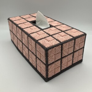 May include: A rectangular tissue box cover constructed from plastic canvas with a grid pattern. The design features light pink yarn stitched in a square motif, outlined by dark charcoal gray yarn borders. A single white tissue protrudes from the top.