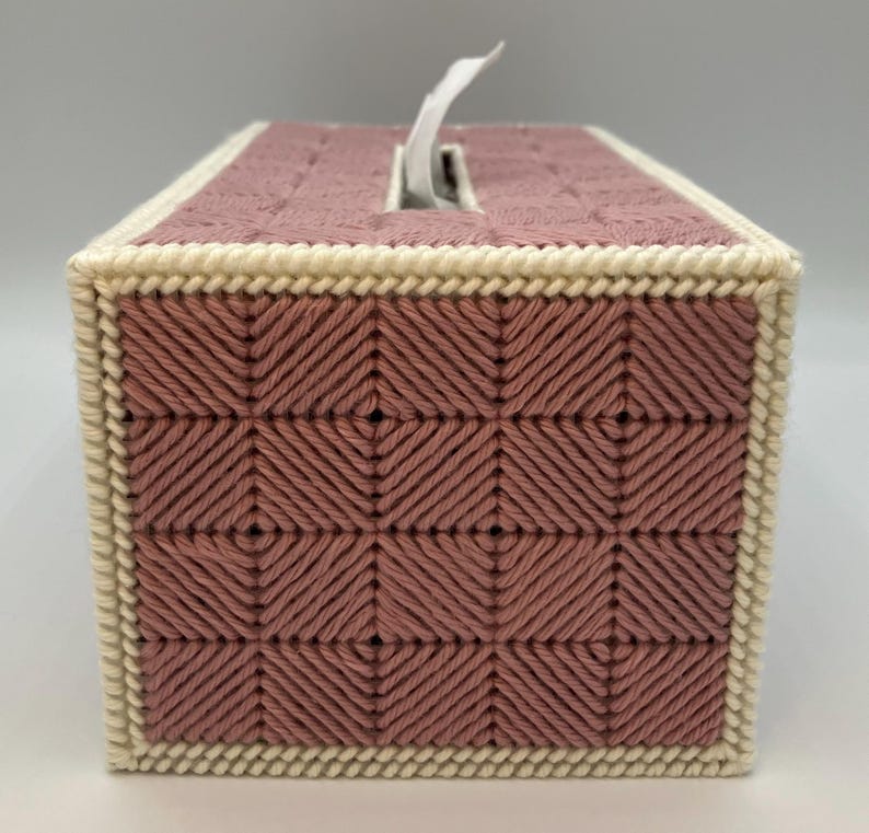 May include: A rectangular tissue box cover in a dusty rose color, featuring a woven pattern. The edges are trimmed with a cream-colored border. A white tissue is partially pulled from the top opening. The cover is handmade.