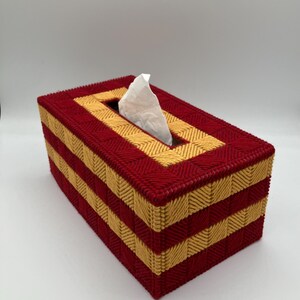 Puede incluir: Una funda rectangular para caja de pa&ntilde;uelos hecha de malla pl&aacute;stica con un patr&oacute;n tejido de hilos alternos en color rojo oscuro y amarillo dorado. La caja presenta una abertura rectangular central en la parte superior con un pa&ntilde;uelo blanco.