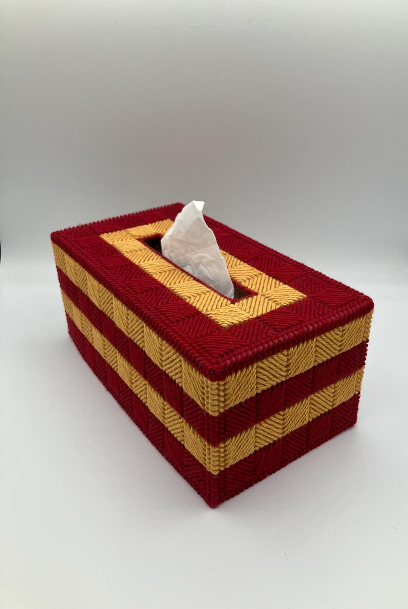 Puede incluir: Una funda rectangular para caja de pa&ntilde;uelos hecha de malla pl&aacute;stica con un patr&oacute;n tejido de hilos alternos en color rojo oscuro y amarillo dorado. La caja presenta una abertura rectangular central en la parte superior con un pa&ntilde;uelo blanco.