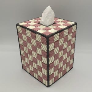 May include: A square tissue box cover with a woven pattern in pink, cream, and dark gray. The design creates a checkerboard effect. A white tissue is visible at the top. The box is a decorative item for home use.