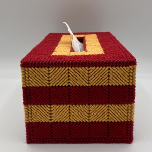 Puede incluir: Una funda rectangular para caja de pa&ntilde;uelos hecha de lona pl&aacute;stica con un patr&oacute;n de hilo cosido. El dise&ntilde;o presenta rayas horizontales alternas de color rojo oscuro y amarillo dorado en punto de espiga, con un pa&ntilde;uelo blanco sobresaliendo.