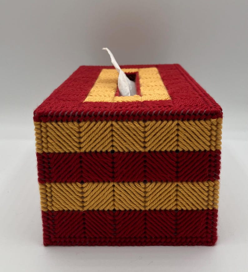 Puede incluir: Una funda rectangular para caja de pa&ntilde;uelos hecha de lona pl&aacute;stica con un patr&oacute;n de hilo cosido. El dise&ntilde;o presenta rayas horizontales alternas de color rojo oscuro y amarillo dorado en punto de espiga, con un pa&ntilde;uelo blanco sobresaliendo.