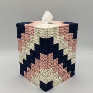May include: A square tissue box cover in a geometric pattern. The cover is made of yarn in navy blue, light pink, and off-white. A white tissue is visible at the top. The box has a woven, textured appearance.