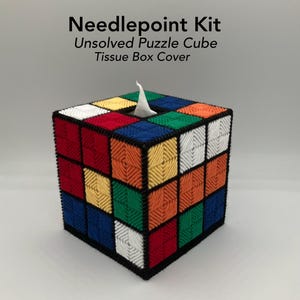 May include: A needlepoint kit for an unsolved puzzle cube tissue box cover. The square cover features a grid of red, blue, green, yellow, orange, and white yarn squares stitched onto a black frame, with a central opening for tissues.