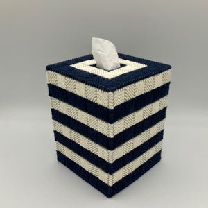 May include: A square tissue box cover with a navy blue and cream striped pattern. The box is made of woven material and has a square opening at the top for tissue dispensing. A white tissue is visible.