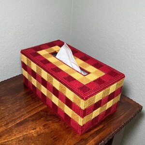 Puede incluir: Una caja para pa&ntilde;uelos rectangular con un dise&ntilde;o de malla pl&aacute;stica tejida en un patr&oacute;n de cuadros que alterna cuadrados de color rojo oscuro y amarillo dorado. Un pa&ntilde;uelo blanco sobresale por la abertura rectangular central.