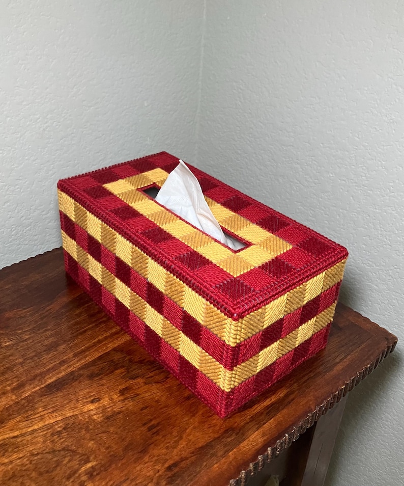 Puede incluir: Una caja para pa&ntilde;uelos rectangular con un dise&ntilde;o de malla pl&aacute;stica tejida en un patr&oacute;n de cuadros que alterna cuadrados de color rojo oscuro y amarillo dorado. Un pa&ntilde;uelo blanco sobresale por la abertura rectangular central.