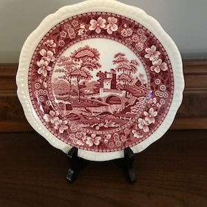 Puede incluir: Un plato de porcelana vintage rojo y blanco con borde festoneado. El plato presenta un borde floral y un diseño central de un puente sobre un arroyo con árboles y una cabaña en el fondo.