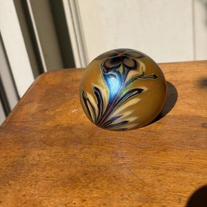 May include: A spherical glass paperweight featuring an iridescent gold and amber base with a swirling floral pattern in shades of deep blue, black, and cream. A small, bright orange dot is centered at the top of the flower design.