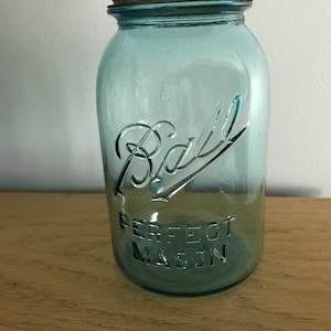 Pode incluir: Um frasco Ball Perfect Mason azul vintage com tampa de metal. O frasco tem letras em relevo e um design clássico, perfeito para conservas ou exibição. O frasco está sobre uma superfície de madeira.