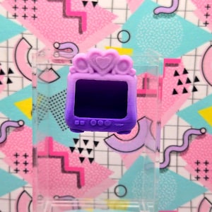 May include: A small, purple toy television with a heart-shaped crown detail. The screen is black, and the base has three circular buttons. The background features a colorful geometric pattern with pink, blue, and yellow shapes.