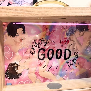 Op de afbeelding: Een houten kunstwerk met een roze achtergrond, met anime-achtige illustraties van mensen. Het kunstwerk bevat de woorden "enjoy GOOD time" en afbeeldingen van vlinders, harten en regenbogen.