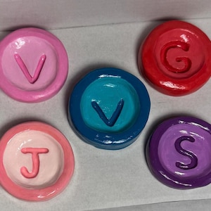 Könnte beinhalten: Eine Sammlung von fünf bunten, runden, bemalten Objekten. Jedes Objekt zeigt einen anderen Buchstaben: V, G, J, S und V. Die Buchstaben sind erhaben und in einer Kontrastfarbe zur Grundfarbe des Objekts gehalten. Die Objekte sind in Rosa-, Rot-, Blau- und Lilatönen gehalten.