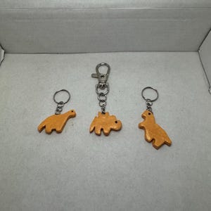 May include: Three orange dinosaur keychains. One keychain has a silver clasp, while the other two have silver key rings. The dinosaurs are different shapes and sizes.