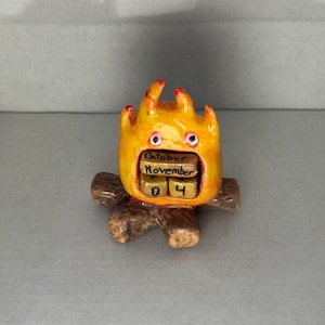 May include: A whimsical, handcrafted figurine of a cartoonish fire with a calendar display. The fire is primarily yellow and orange with red accents. The calendar shows "October" and "November" with the date "04". The base is made of brown wooden logs.