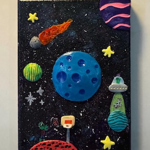 May include: A whimsical, handmade space-themed artwork on canvas. The painting features a blue planet, a green and yellow striped sphere, a red comet, yellow stars, and a green and blue UFO. A red planet with a basketball hoop and two aliens is also depicted.