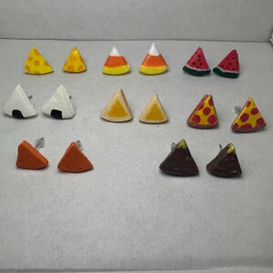 May include: A collection of handmade stud earrings in various food shapes. Designs include cheese, candy corn, watermelon, onigiri, pizza, and more. Each earring is a small, colorful triangle with a post for pierced ears. The earrings are arranged on a white surface.