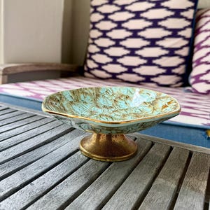 May include: A shallow, footed bowl featuring a textured, light turquoise interior with brown veining and a gold-toned rim. The base is a solid, metallic gold pedestal that supports the wide, slightly irregular, and rounded bowl shape.