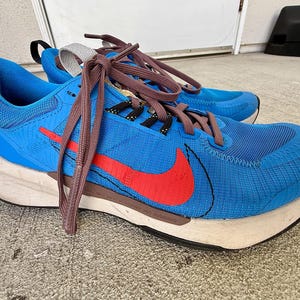 May include: A bright blue athletic sneaker featuring a prominent red swoosh logo on the side, dark brown laces, and a thick, off-white foam midsole. The shoe has a textured mesh upper, a black heel tab, and a grey pull loop on the tongue.