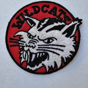 May include: A circular embroidered patch featuring a white wildcat head with black details on a red background. The word WILDCATS is stitched in black capital letters along the top edge of the patch, which has a thick black border.