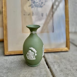 May include: A small, olive green vase with white relief detailing. The vase features a classical design with figures and floral accents. The vase is displayed in front of a framed picture with a gold frame.