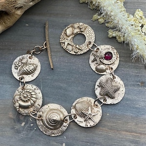Peut inclure: Un bracelet doré composé de sept maillons circulaires, chacun orné de motifs de vie marine en relief, notamment des coquillages et des étoiles de mer. Un maillon contient une pierre précieuse ronde et facettée rose foncé. Fermoir à barre.
